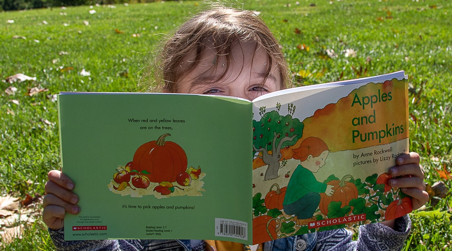 A young girl with dark blonde hair is outside sitting in the grass, with a book entitled Apples and Pumpkins, and she peeks over the book to look at the camera
