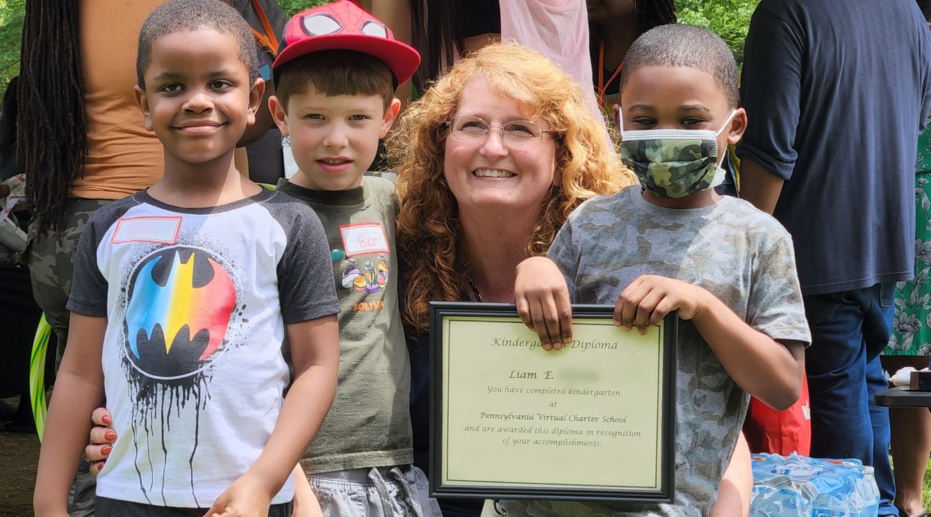 A Kindergarten teacher is surrounded by a diverse group of three male kindergartners, and one student of color is holding his diploma