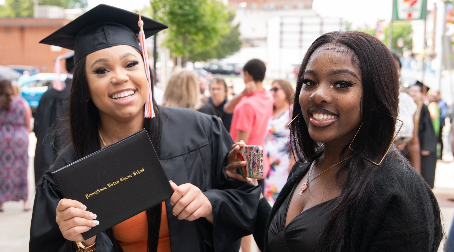 A 2022 female graduate of color is in her cap and gown holds up her PA Virtual Charter School diploma, and smiles at the camera, while her friend holds a phone up and also smiles