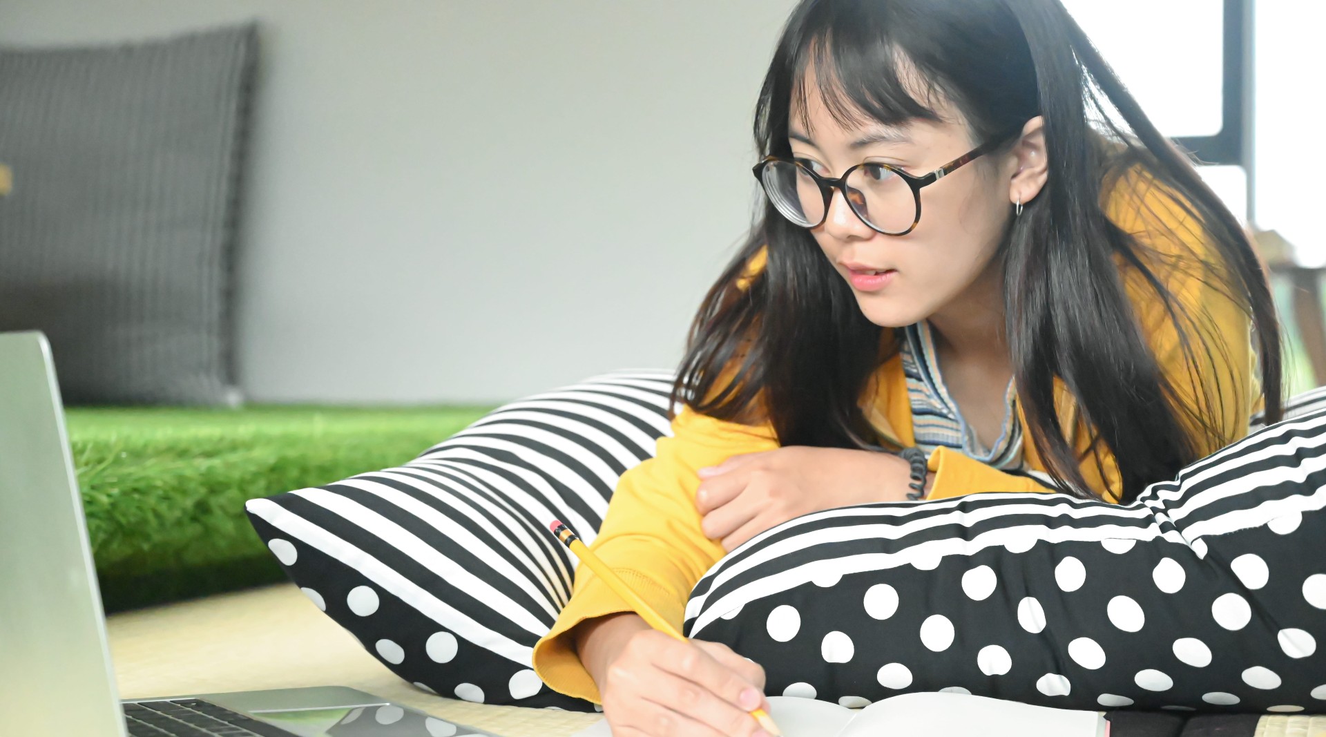 A young Asian woman with glasses is in a yellow sweater and is laying on her stomach on a black and white striped and polka dot pillow, as she works on her computer