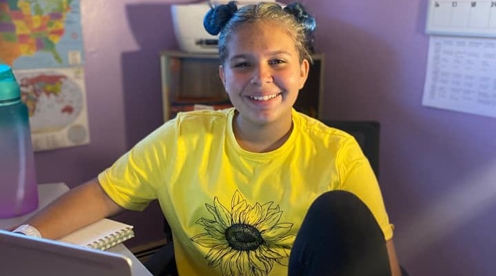 Blond female seventh grader smiles at camera while working on her computer at her desk at home