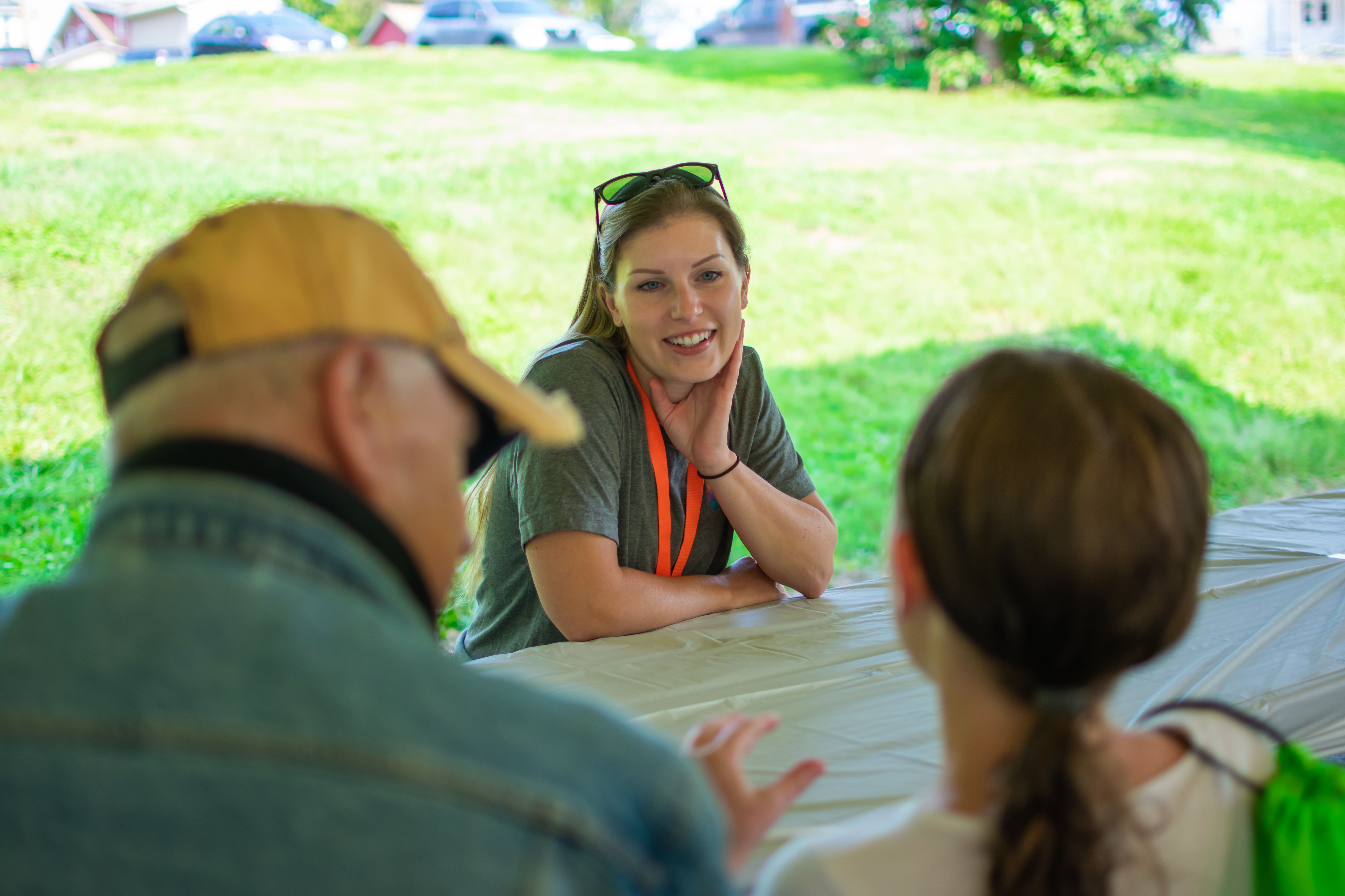 A smiling blond female PA Virtual teacher is wearing an orange lanyard and has sunglasses on her head, and is listening to a young girl and her grandfather speak. We only see the back of the heads of the girl and grandfather. All three are outside.