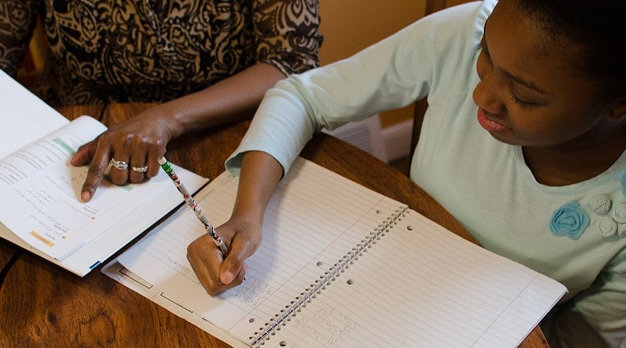 young student of color works at table, writing on a notebook with her pencil, while a female adult sits beside her and points to a page in the textbook